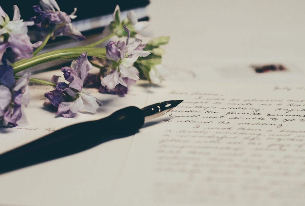 a piece of paper with cursive handwriting and an ink pen, with purple flowers lying on the paper