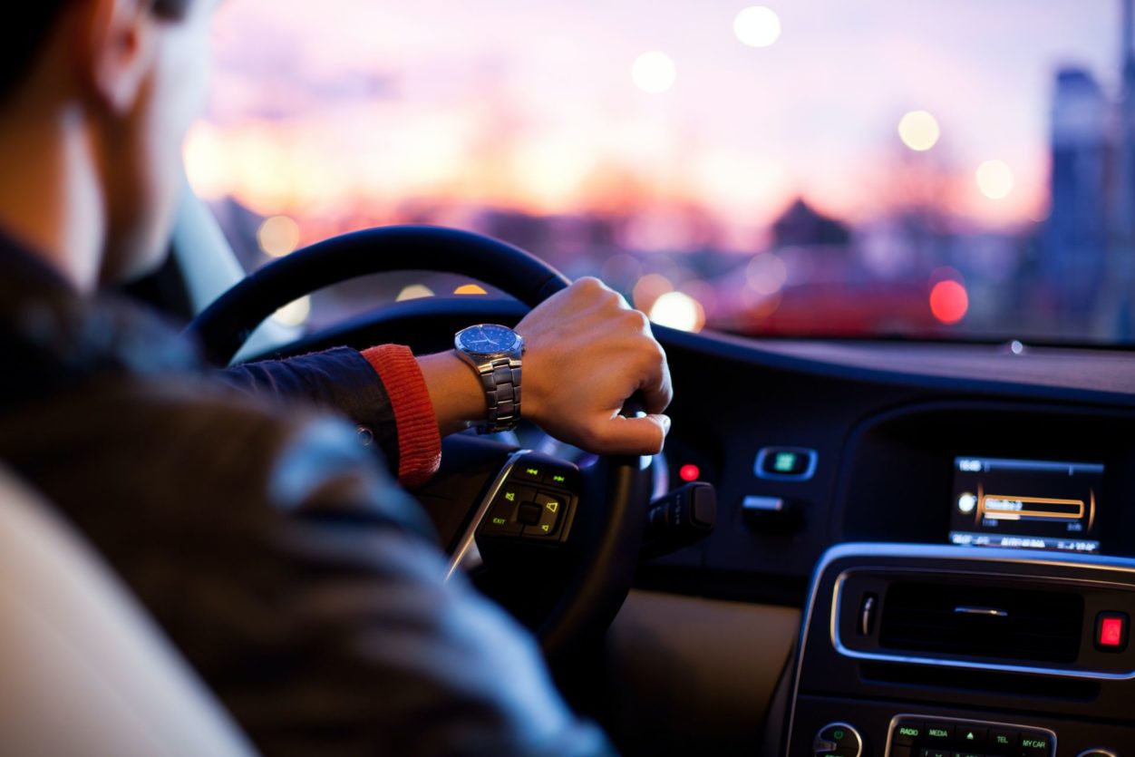 closeup of a mans hand holding a steering wheel from behind with sunset city outside window