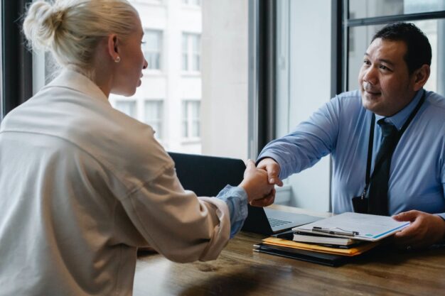 a job interview highlighting the new changes to the pay transparency act; a woman shaking hands with a man across the desk, holding a clipboard with interview questions on it