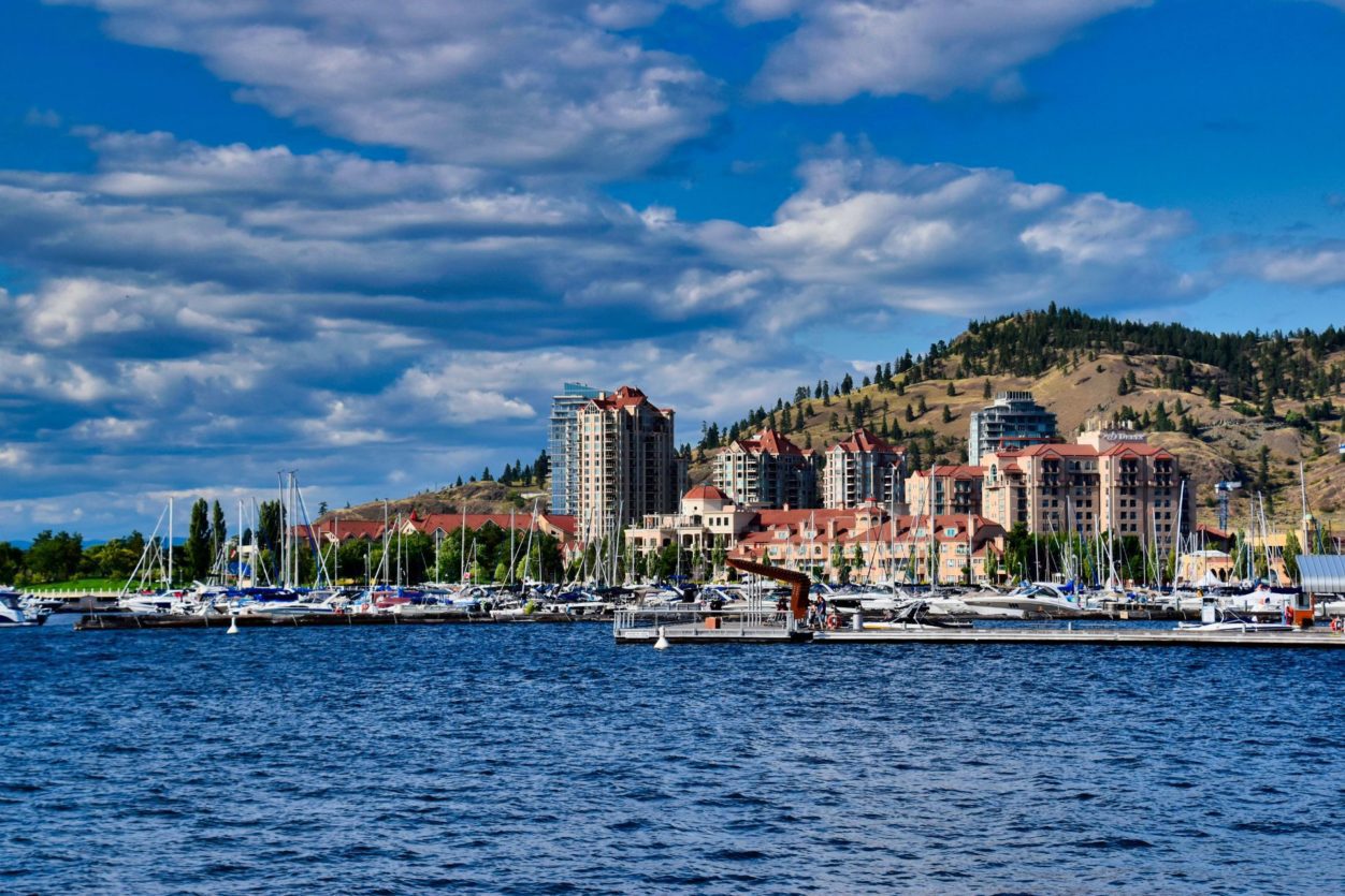 a view of the Kelowna Grand hotels and Yacht Club marina over the water, with mountains and blue skies in the background