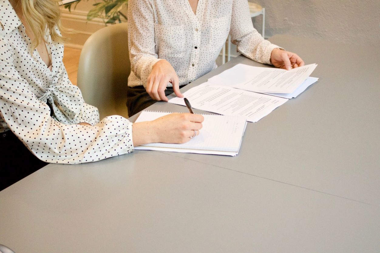 two women in white spotted blouses sitting at a grey table signing paperwork for a mediator contract