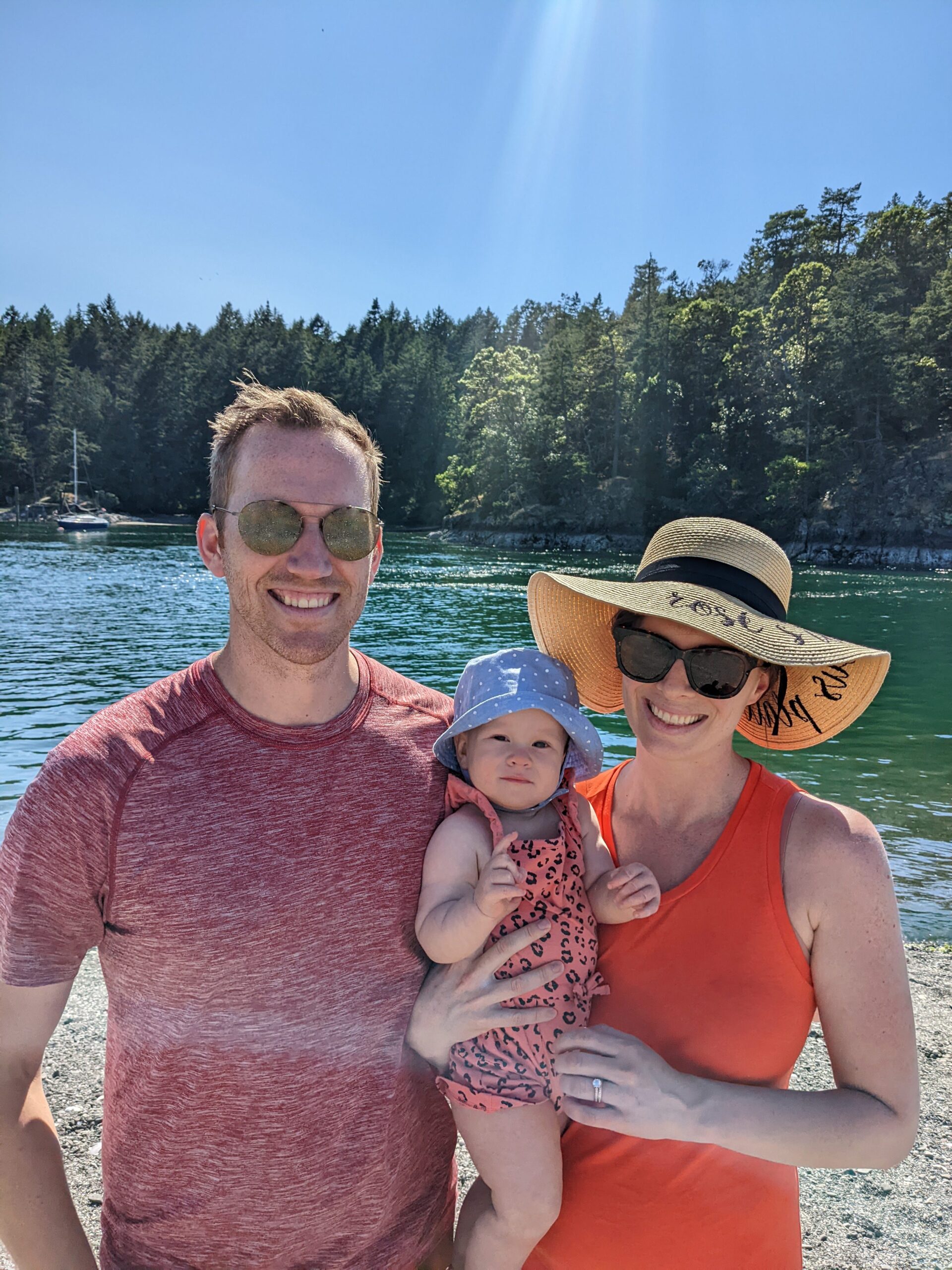 Casual photo of Rosie Jacobs, a brunette in a straw sun hat and orange tank top, holding her baby in a blue hat and pink leopard print onesie, standing next to her husband at the beach.